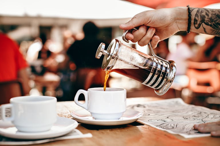 Barista pouring coffee from a French press into a cup on a wooden table in a bustling café.