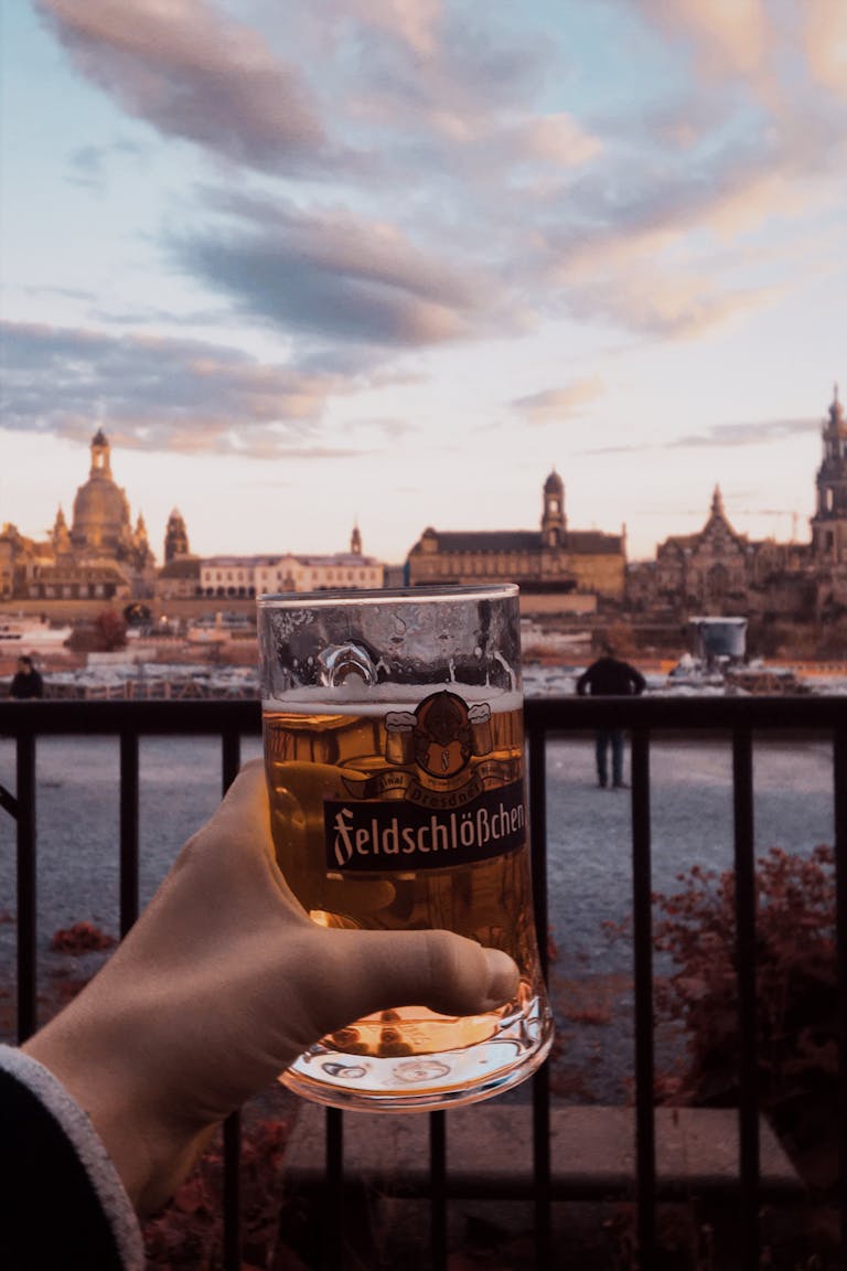 A hand holding a beer glass against the backdrop of a historic cityscape during sunset.