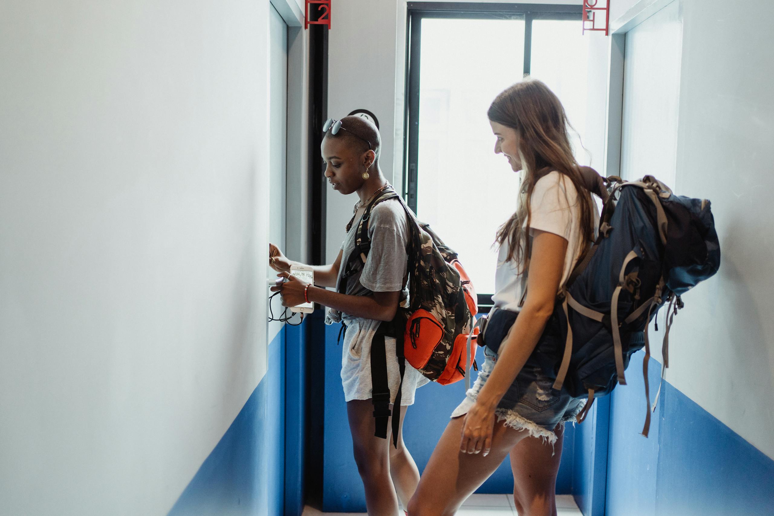 Two women travelers in a hostel corridor with backpacks, preparing for their journey.