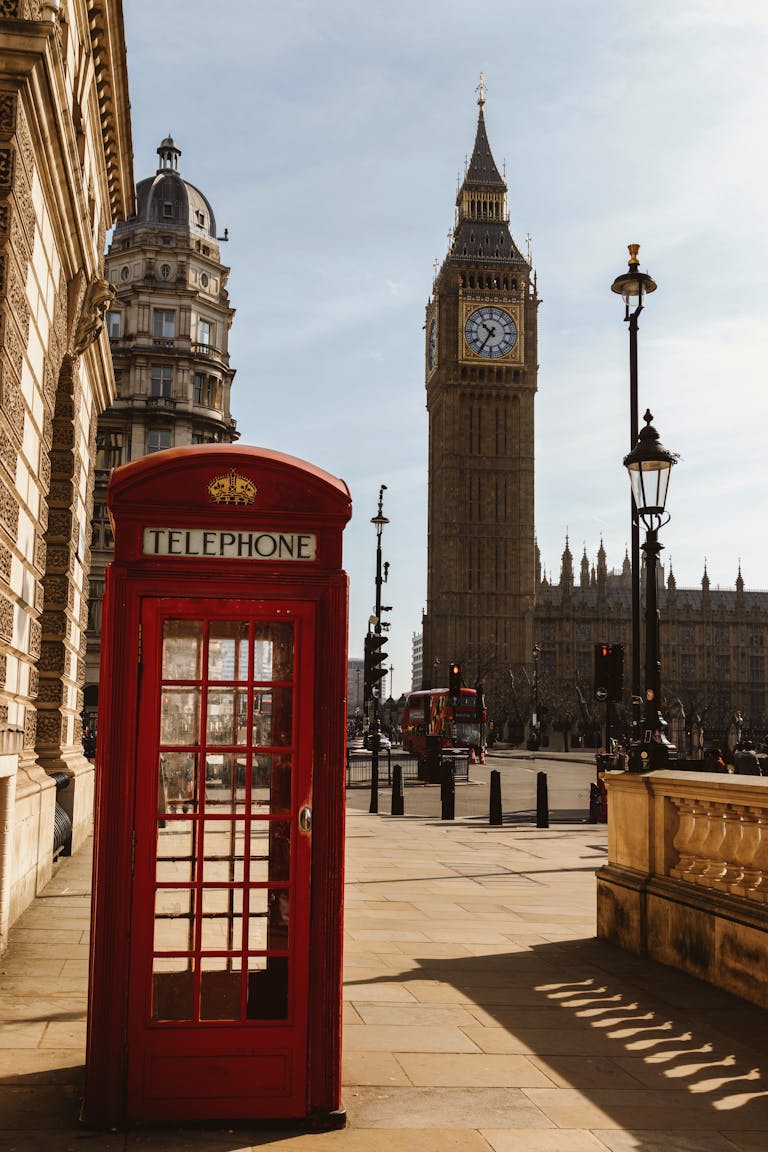 Red telephone booth with Big Ben and historic London architecture in daylight.